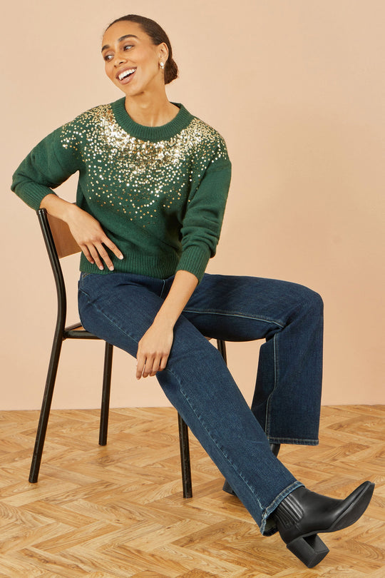 Woman wearing green relaxed knitted jumper with scattered sequin neckline sitting on chair