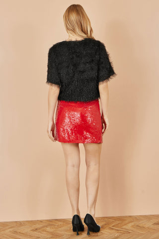 Woman wearing red sequin mini skirt with black textured top and black heels, standing on wooden floor against beige background